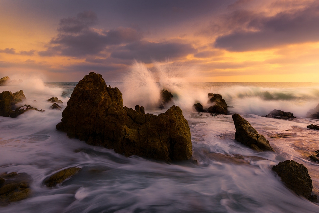 A photograph of Little Corona Beach in California. The waves are hitting the rocks and splashing into the air at sunset. A nature photography print by Bryce Mironuck